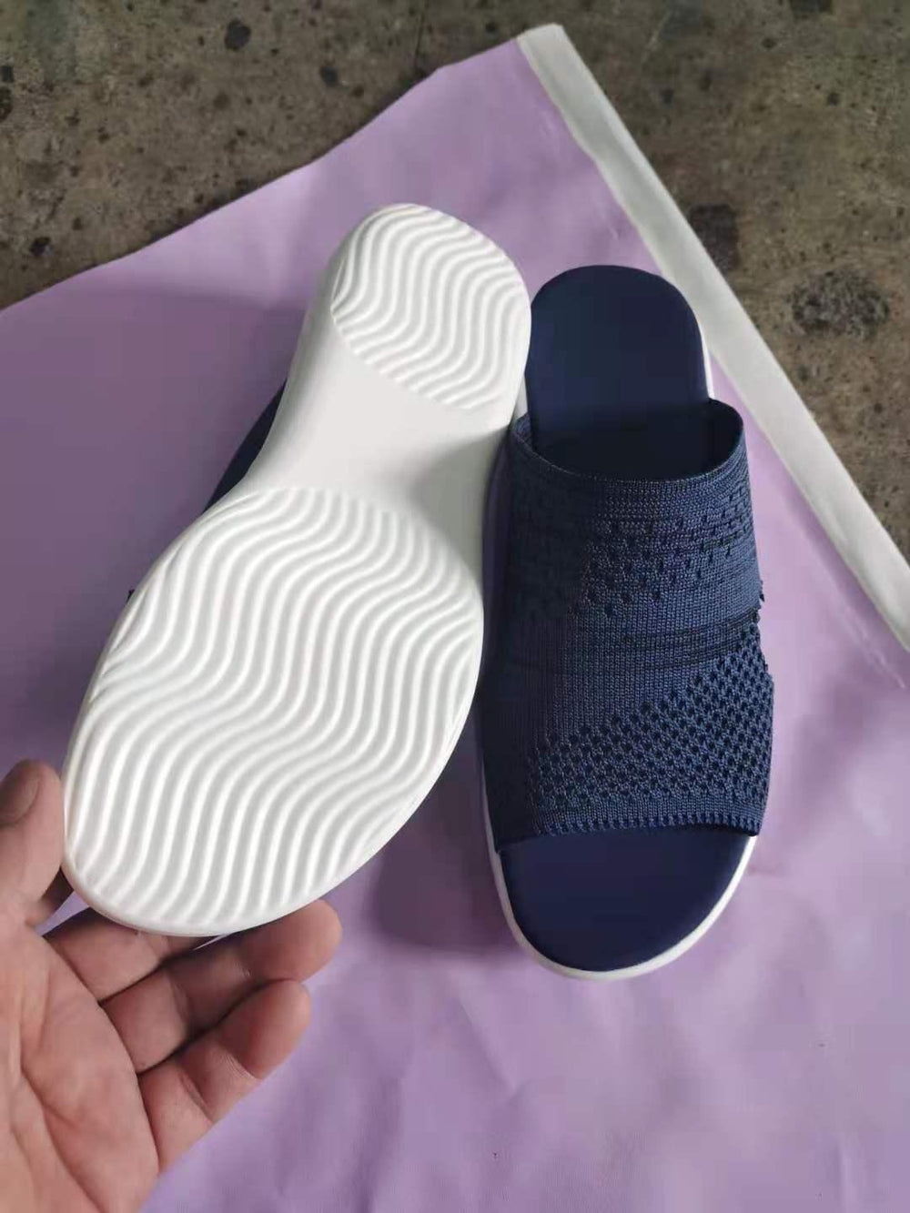Hand holding navy blue slide sandal with white textured sole on purple and gray background