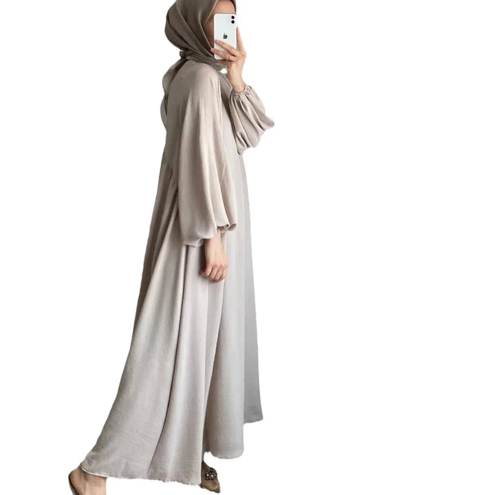Woman wearing a loose beige abaya with balloon sleeves and khaki hijab, taking a mirror selfie