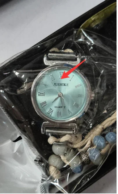 Close-up of a silver NUSHIKE quartz wristwatch with Roman numerals and beaded rope strap in packaging