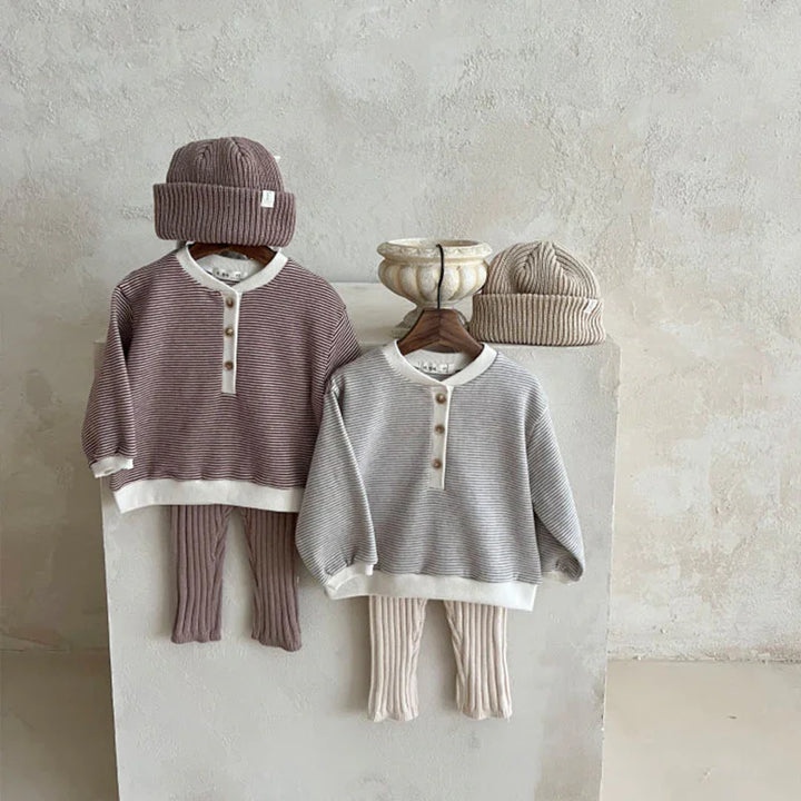 Two toddler outfits with striped long-sleeve tops, ribbed pants, and matching knit beanies in beige and mauve tones displayed on hangers