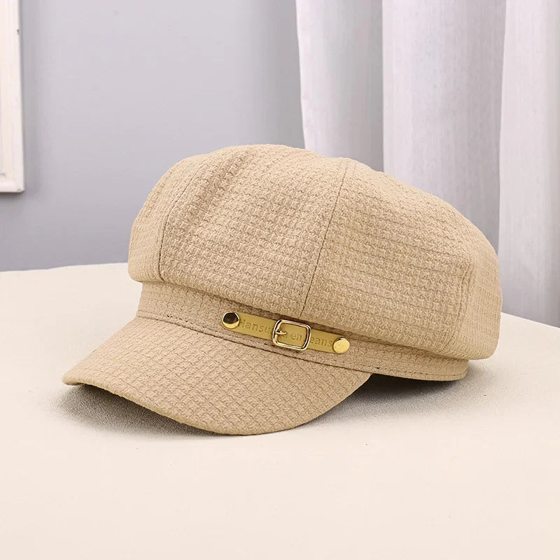 Beige textured fabric newsboy cap with gold buckle detail on beige surface