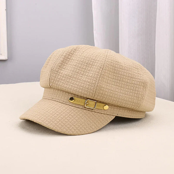 Beige textured fabric newsboy cap with gold buckle detail on beige surface