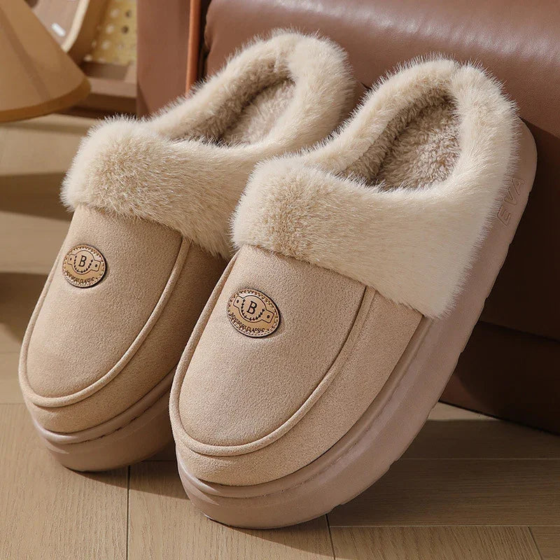 Beige slip-on slippers with plush fur lining and a decorative B emblem on wooden floor