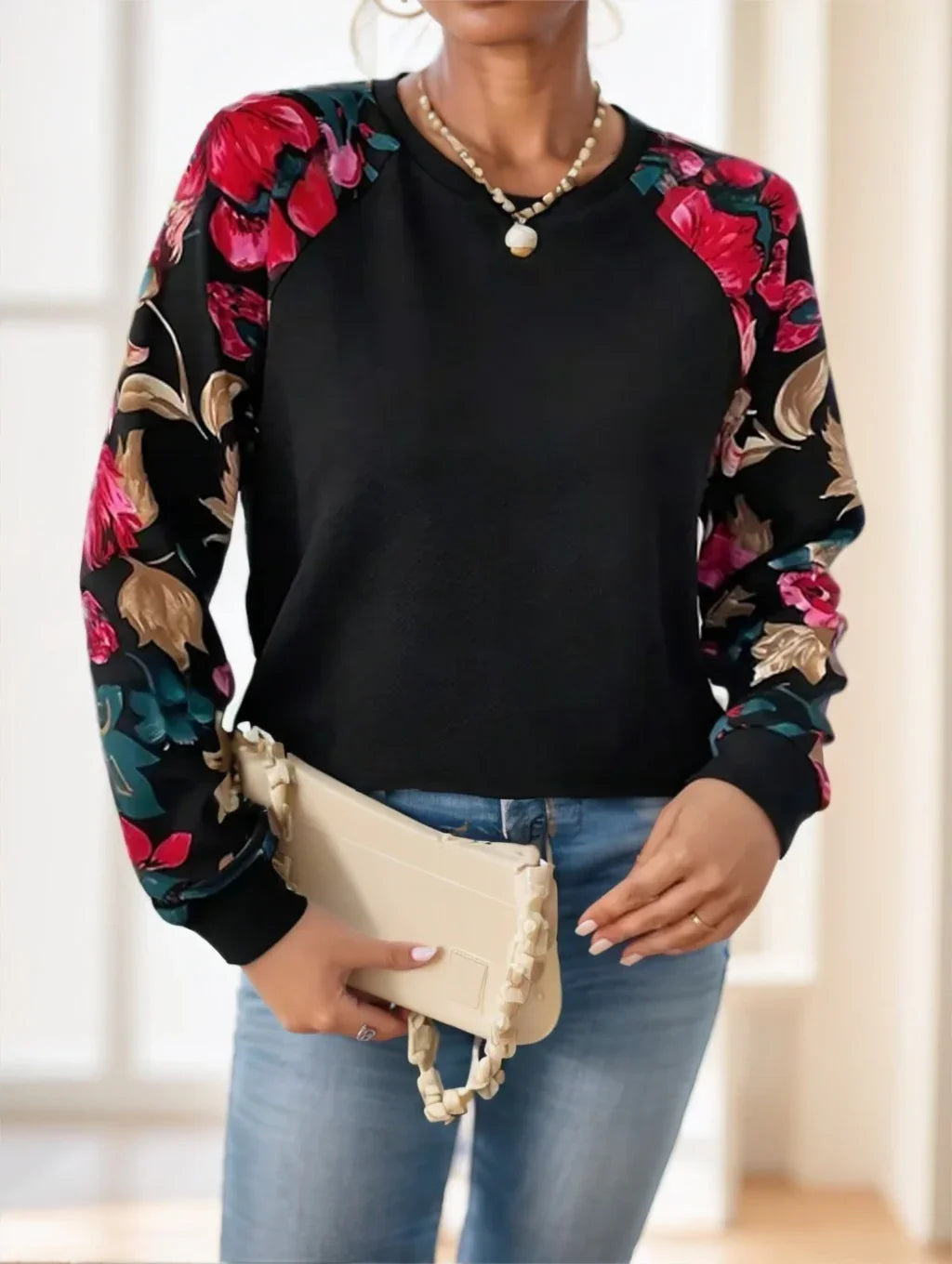 Woman wearing black sweatshirt with colorful floral print sleeves, holding beige handbag, blue jeans