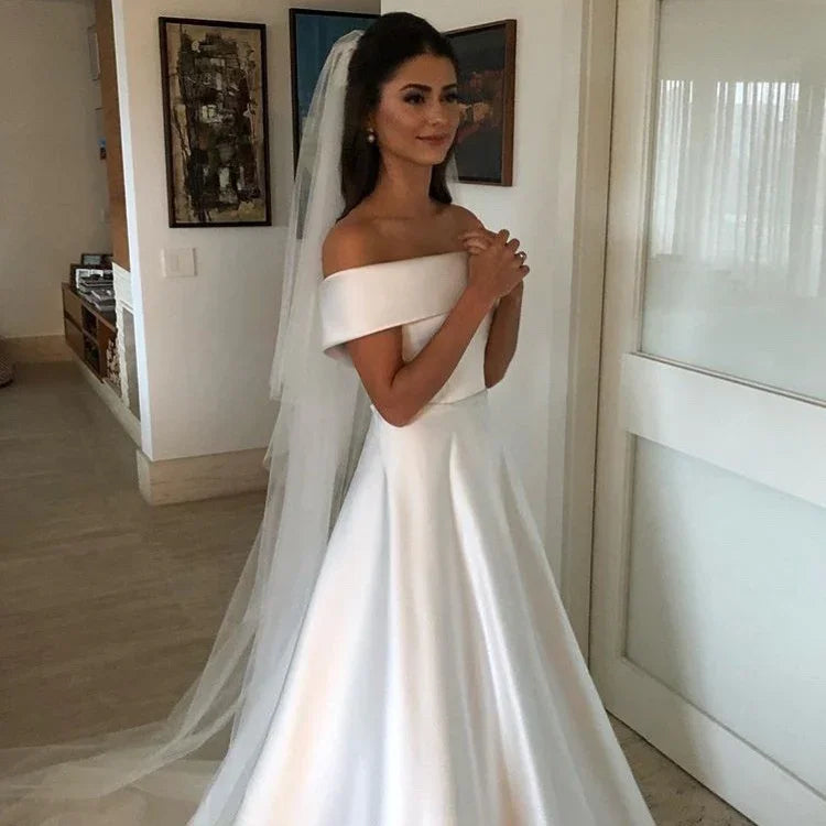 Bride in elegant off-shoulder white satin wedding gown with long veil indoors
