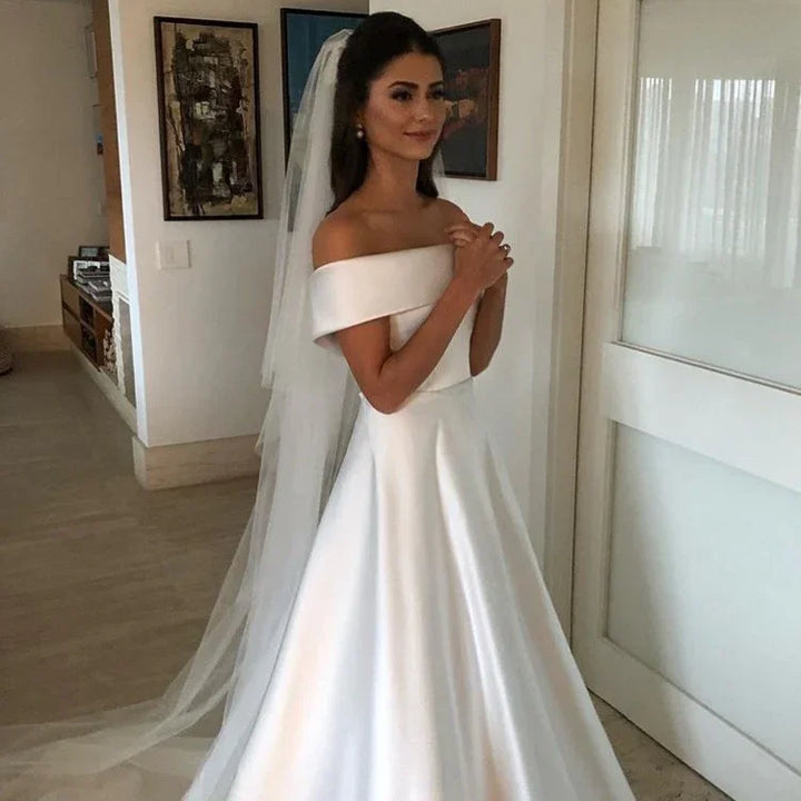 Bride in elegant off-shoulder white satin wedding gown with long veil indoors