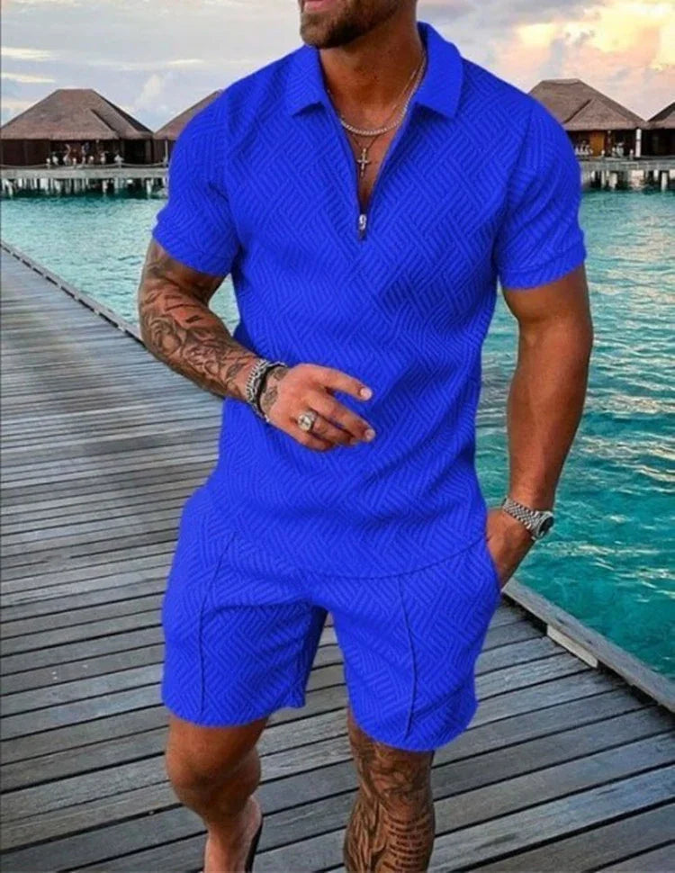 Man wearing blue patterned short-sleeve polo shirt and matching shorts standing on wooden pier over turquoise water with overwater bungalows