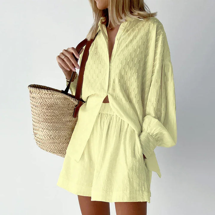 Woman wearing light yellow textured button-up shirt and matching shorts holding woven straw tote bag