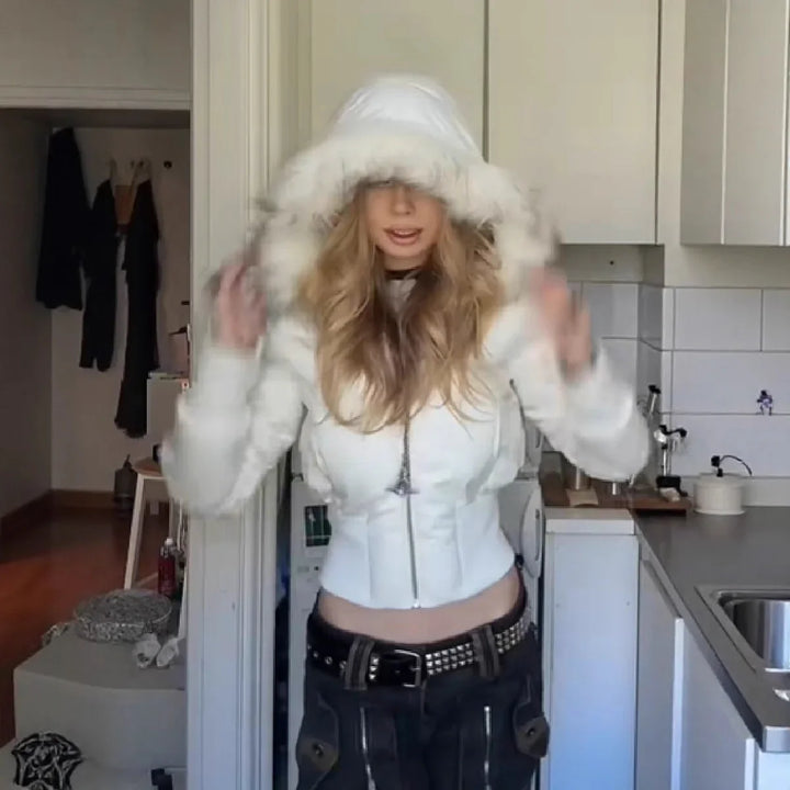 Woman in white fur-trimmed hooded jacket and black pants posing indoors in a modern kitchen