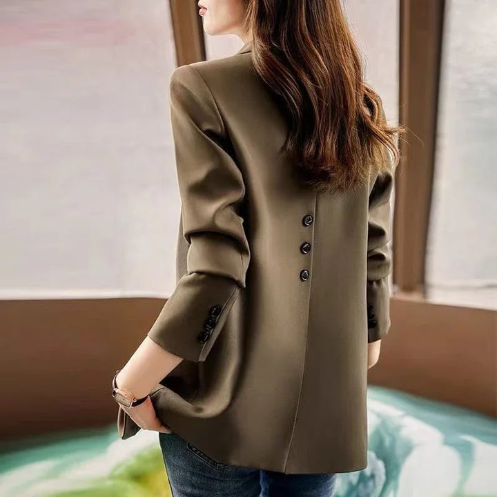 Woman wearing a stylish olive green blazer with buttons on the back, paired with blue jeans indoors