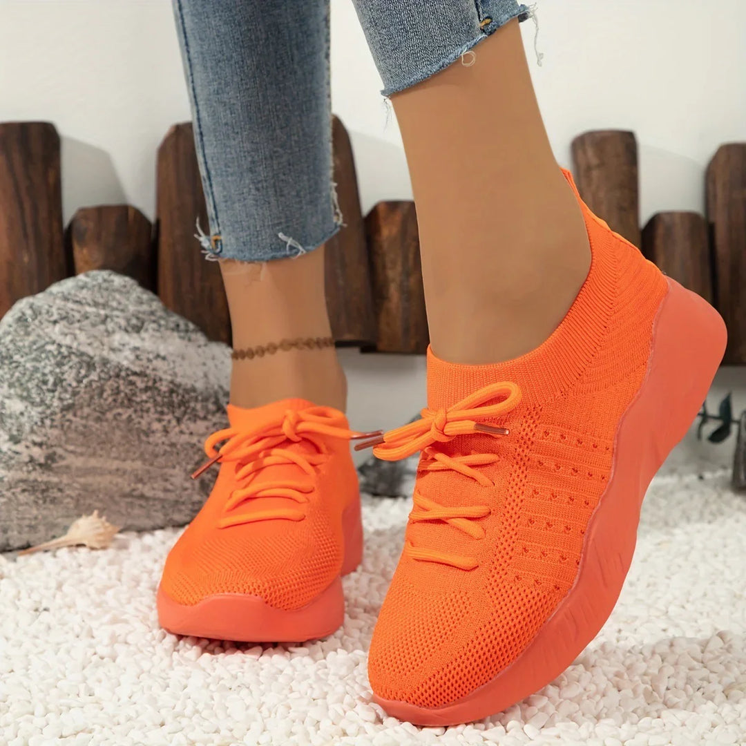 Bright orange knitted sneakers with laces worn with cropped blue jeans on white pebble floor