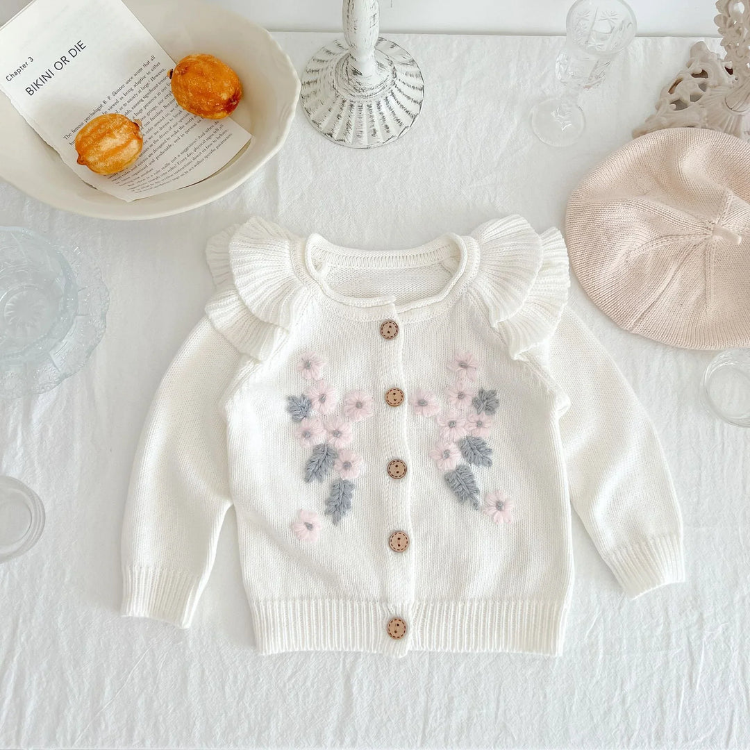 White baby cardigan with pink and gray flower embroidery and ruffled shoulders on a white tablecloth