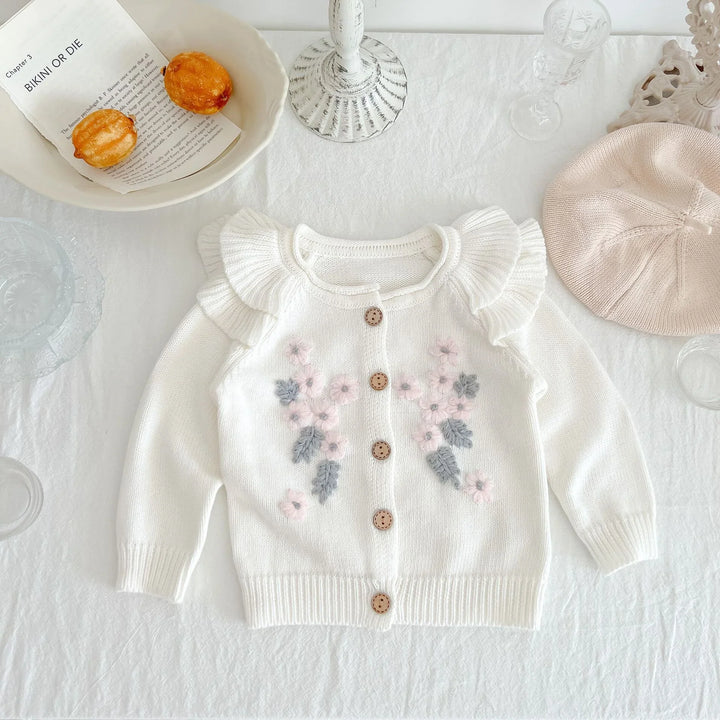 White baby cardigan with pink and gray flower embroidery and ruffled shoulders on a white tablecloth