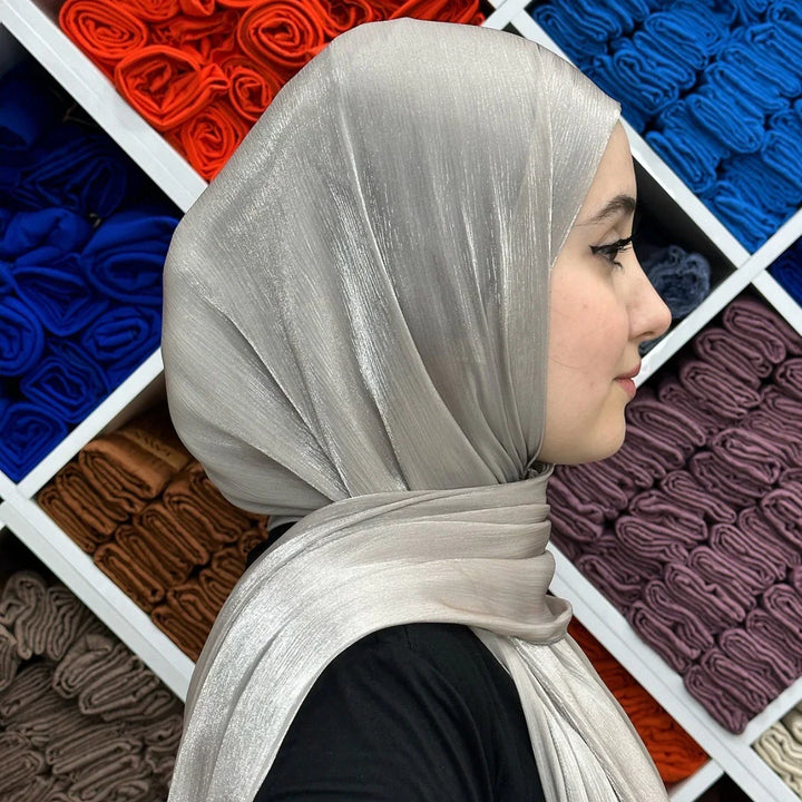Profile of woman wearing shiny silver hijab with colorful folded scarves in the background
