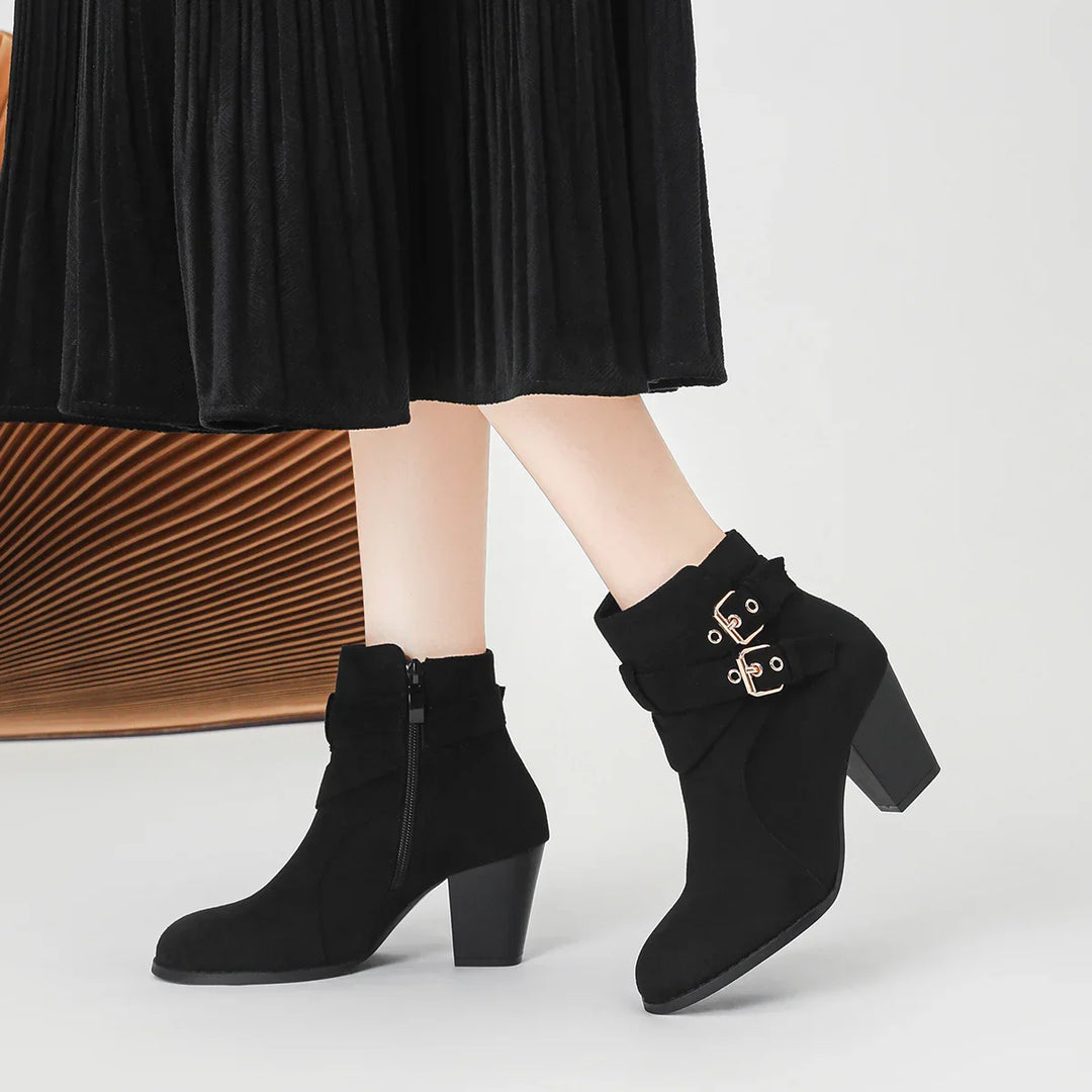 Black suede ankle boots with block heels and gold buckles worn with a black pleated skirt