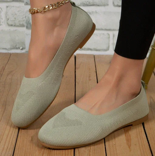 Beige textured women's ballet flats with a gold ankle chain and black leggings on wooden floor