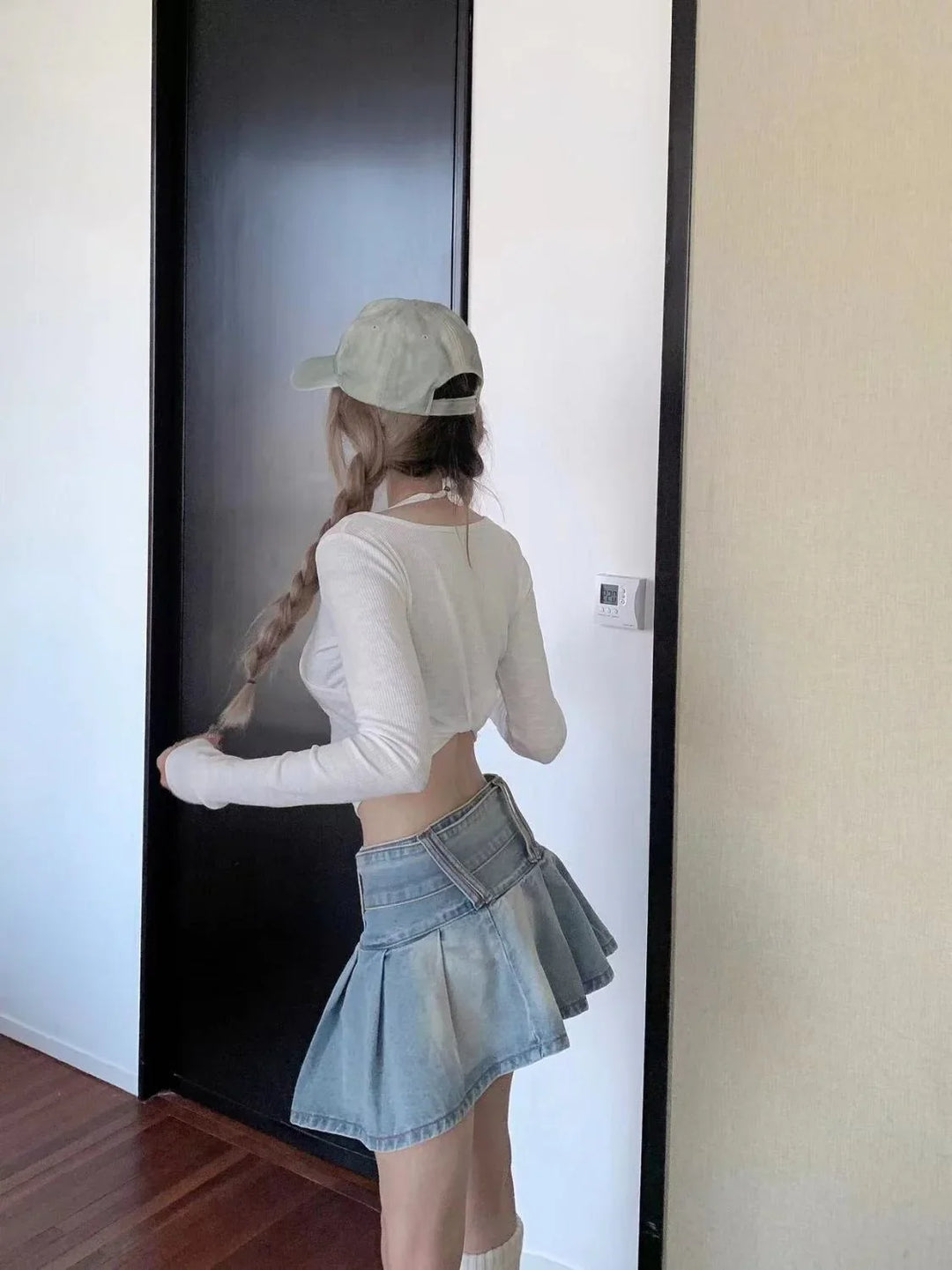 Woman with long braided hair wearing a white crop top, denim pleated skirt, and gray cap indoors