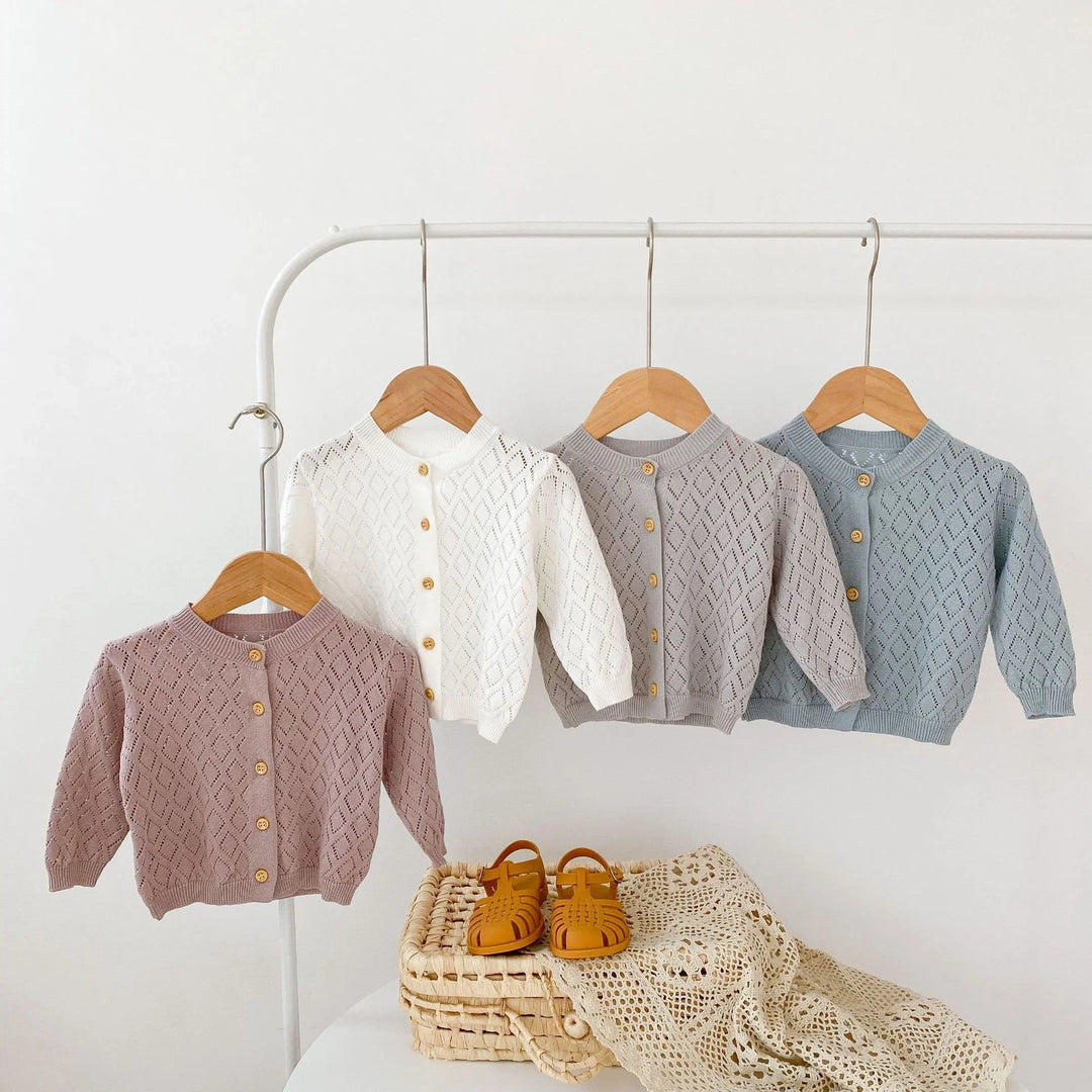 Four baby knitted cardigans in pink, white, beige, and blue on wooden hangers with brown shoes on wicker basket