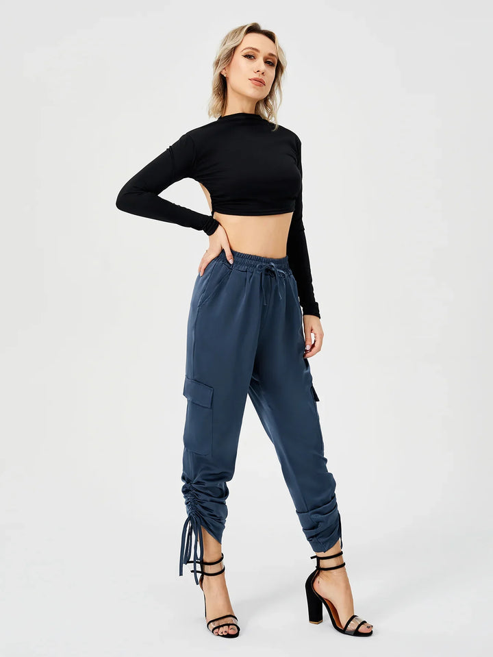 Woman in black long-sleeve crop top and blue ruched cargo pants posing in black high heel sandals on white background
