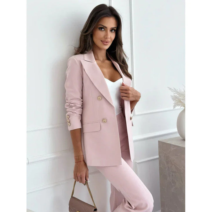 Woman wearing a stylish blush pink suit with gold buttons, holding a taupe handbag in a white room