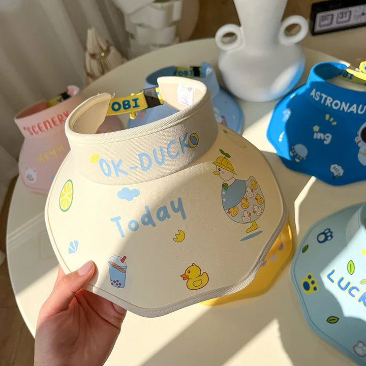 Hand holding beige sun visor with cute duck and boba tea graphics, surrounded by colorful visors on white table