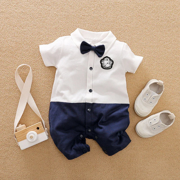 Baby boy two-tone white and navy romper with bow tie, white baby shoes, and toy camera on beige carpet