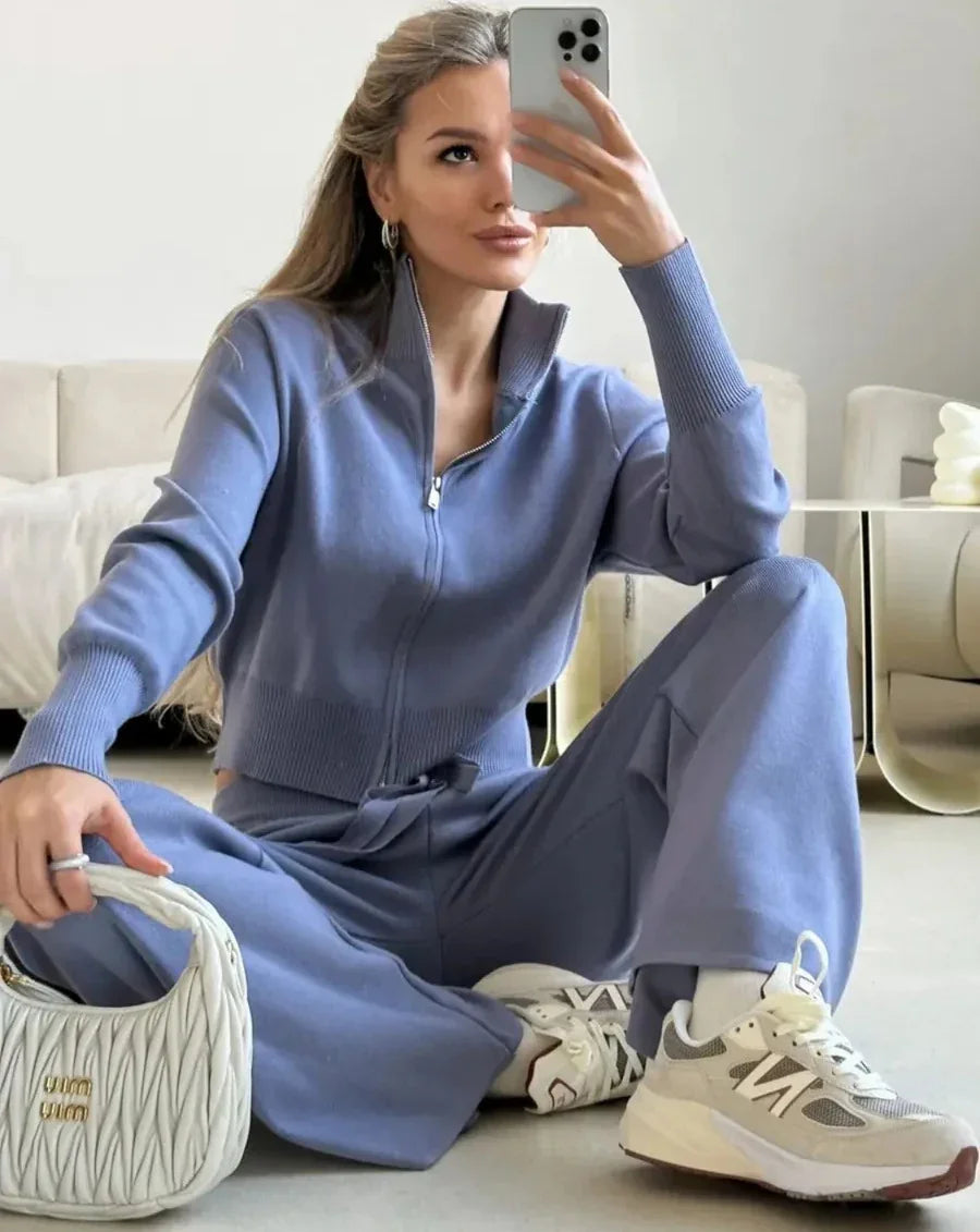 Woman in blue zip-up sweater and matching pants taking mirror selfie wearing white sneakers and holding white purse
