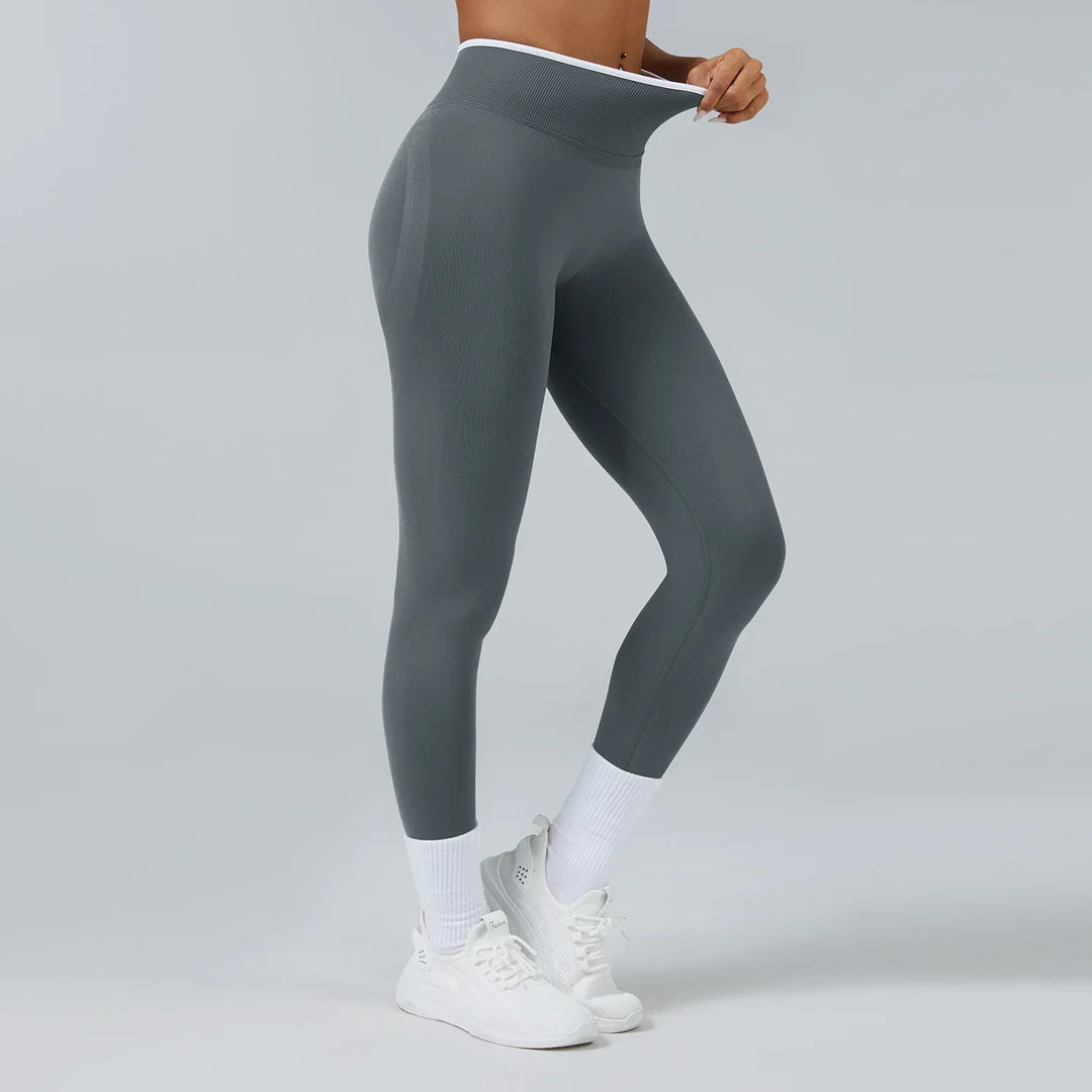 Woman wearing high-waisted grey seamless leggings with white trim and white sneakers on grey background