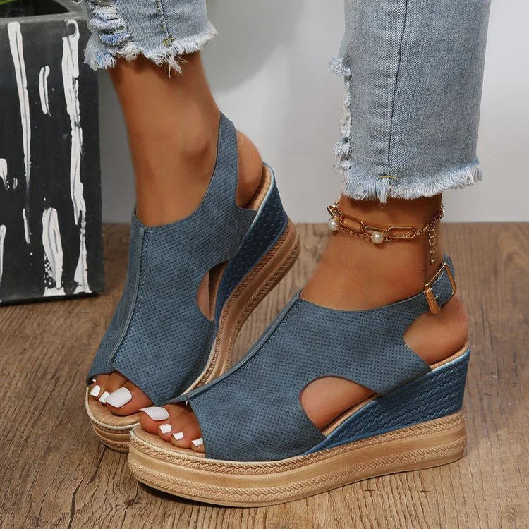 Blue open-toe wedge sandals with ankle strap on feet wearing distressed light blue jeans and gold ankle bracelet