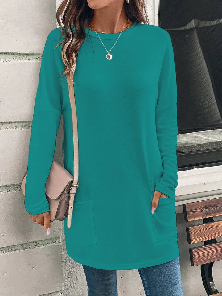 Woman wearing teal long sleeve tunic with front pockets, carrying beige crossbody bag, standing by bench