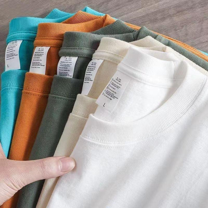 Hand holding a stack of folded solid color cotton t-shirts in white, beige, green, orange, and blue