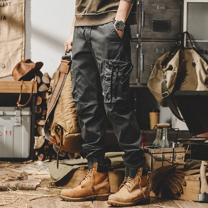 Man wearing black cargo pants and tan work boots holding a brown backpack in rustic workshop
