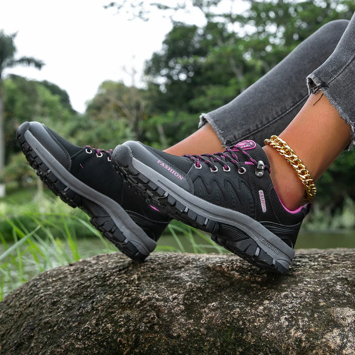Black and gray outdoor hiking shoes with pink accents worn with rolled-up jeans and gold anklet on rock