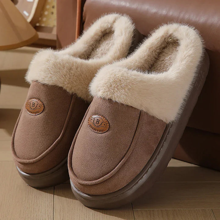 Brown suede slippers with plush beige fur lining and cushioned sole on wooden floor