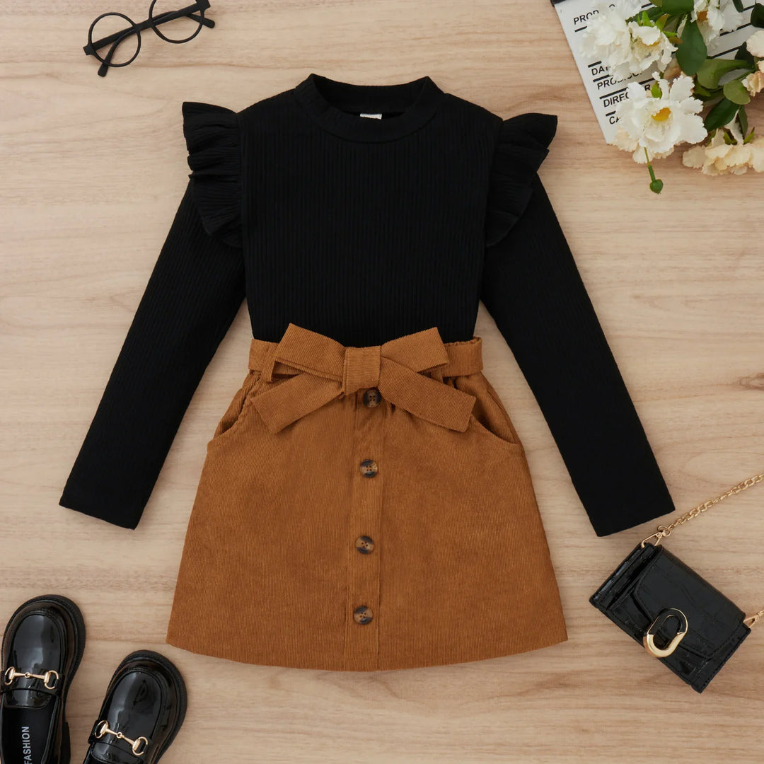 Black long-sleeve top with ruffle shoulders paired with a brown button-front skirt with bow belt on wooden floor