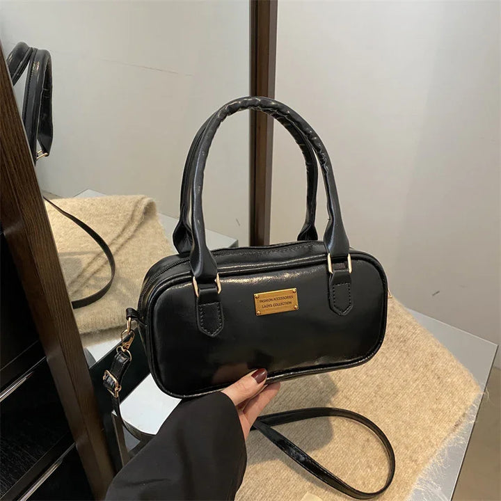 Black leather handbag with gold label reading Fashion Accessories Lady's Collection, shown with hand and beige cloth background