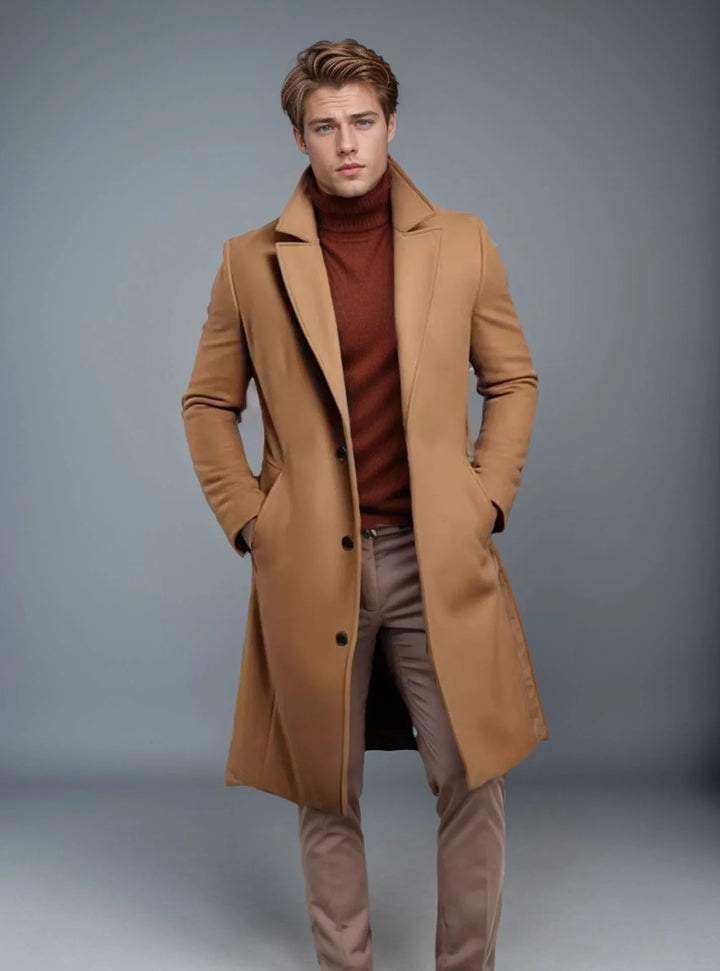 Young man wearing a camel overcoat, rust turtleneck sweater, and beige pants against gray background