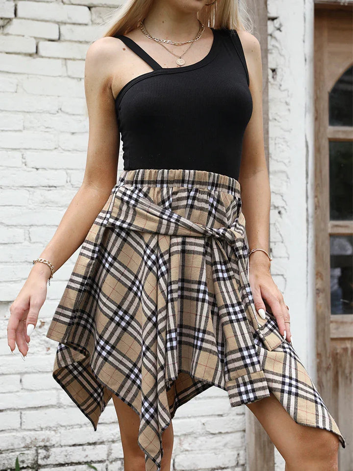 Woman wearing black one-shoulder top and beige plaid asymmetrical skirt standing by white brick wall