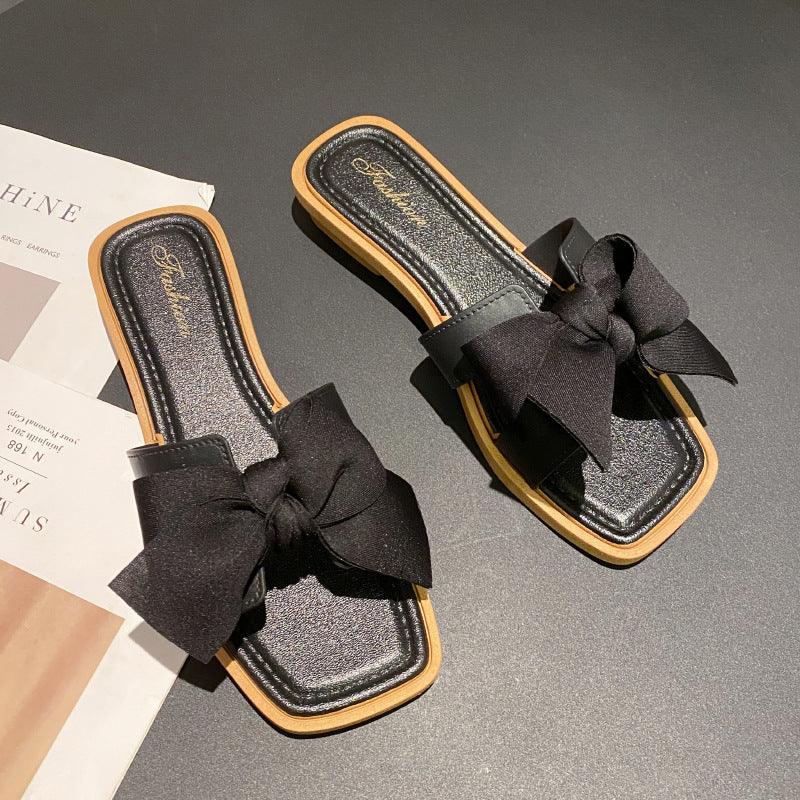 Black slide sandals with large fabric bows and tan edges on black surface next to magazine
