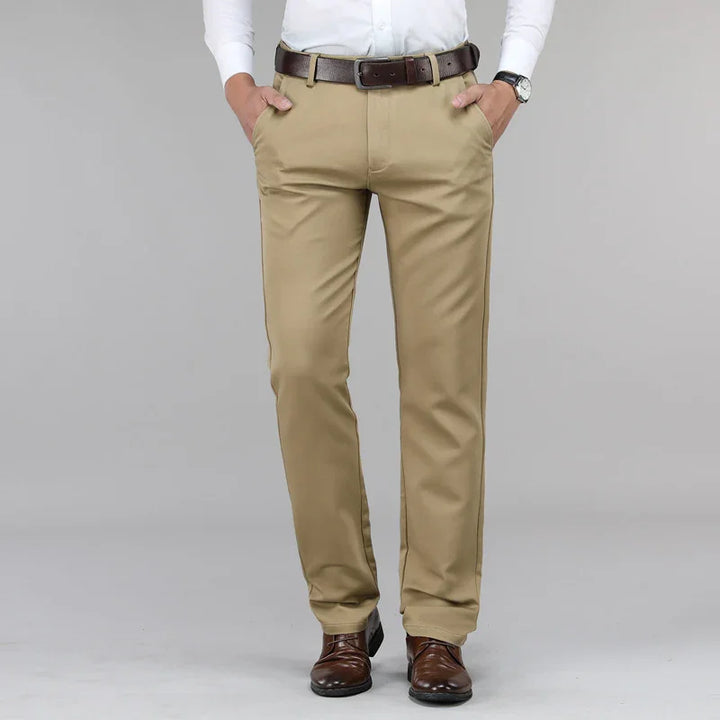 Man wearing beige dress pants with brown leather belt, white shirt, and brown dress shoes on gray background