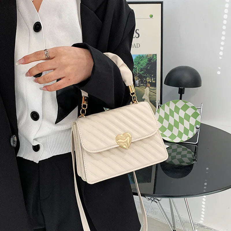 Woman in black blazer holding small cream quilted handbag with gold heart clasp in modern room