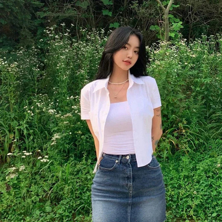 Young woman in white shirt and denim skirt posing outdoors with green plants and white flowers