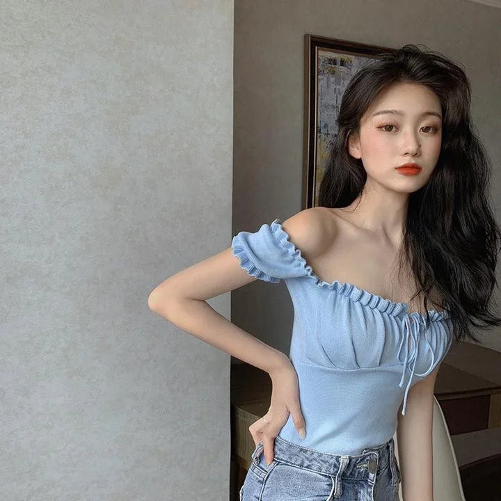 Young woman with long black hair wearing a light blue off-shoulder top and denim jeans indoors