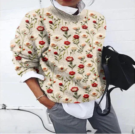 Woman wearing beige sweater with red and orange floral embroidery layered over white shirt, gray pants, and holding black bag