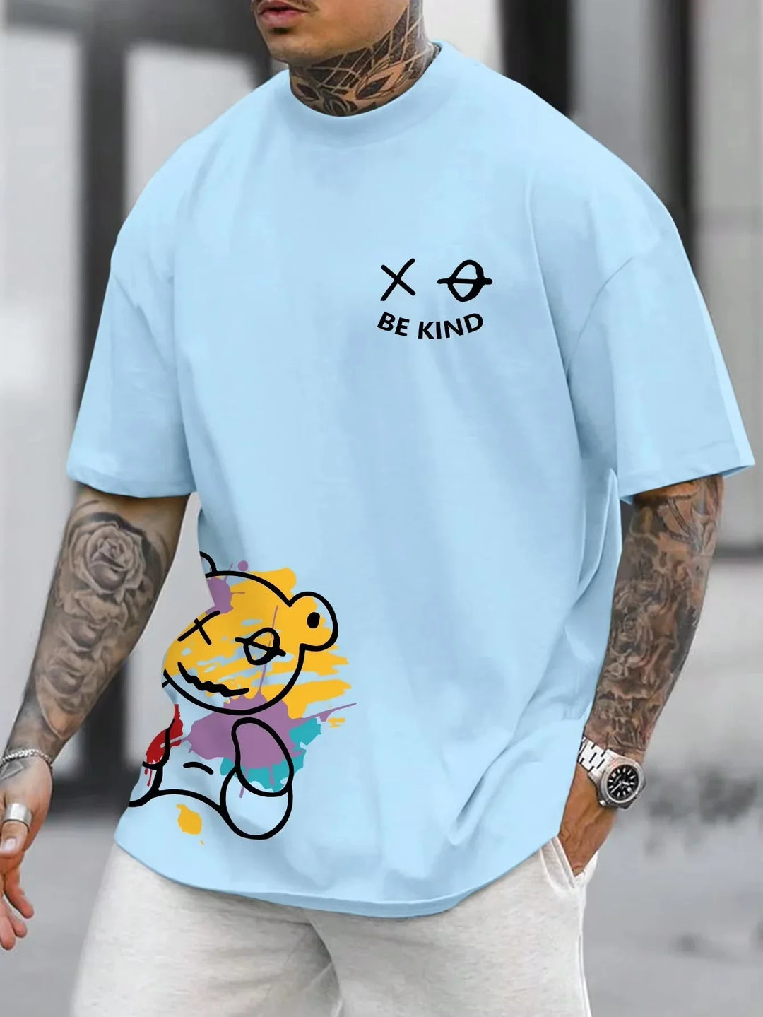 Man wearing light blue oversized t-shirt with 'Be Kind' text and colorful abstract teddy bear graphic
