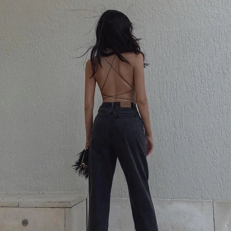 Woman wearing backless strappy top with high-waisted black jeans holding fringe handbag against textured wall