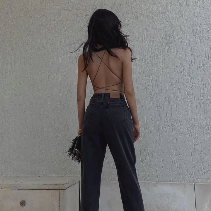 Woman wearing backless strappy top with high-waisted black jeans holding fringe handbag against textured wall