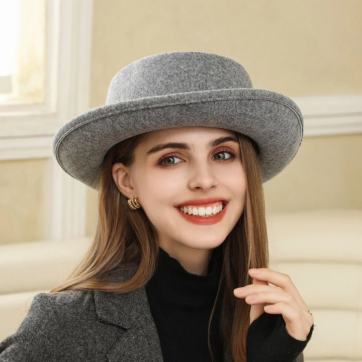 Smiling woman wearing a gray wool hat, gold hoop earrings, black turtleneck, and gray coat indoors