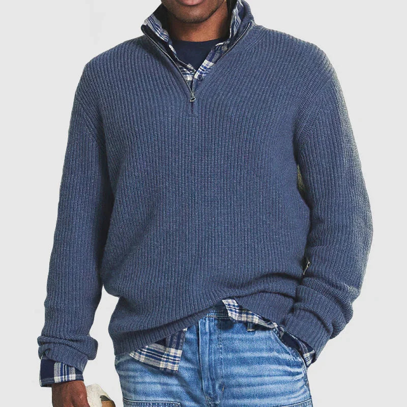 Man wearing blue quarter-zip sweater over plaid shirt with blue jeans on white background
