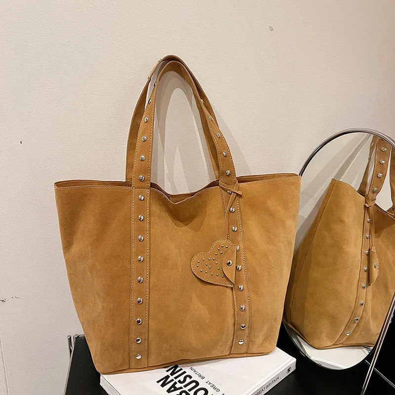 Tan suede tote bag with silver studs and heart charm, displayed on white book with mirror reflection